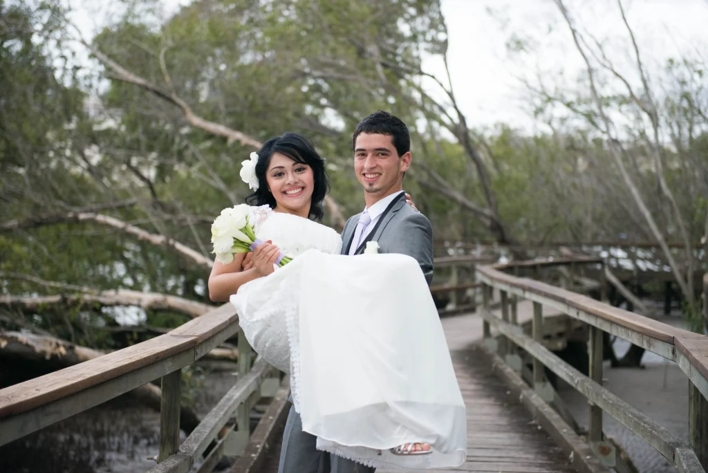 The groom carries the smiling bride on a tree-lined boardwalk.