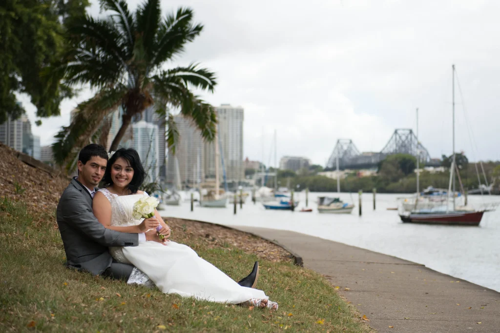The couple sit beside the harbour, with boats and a bridge in the distance.