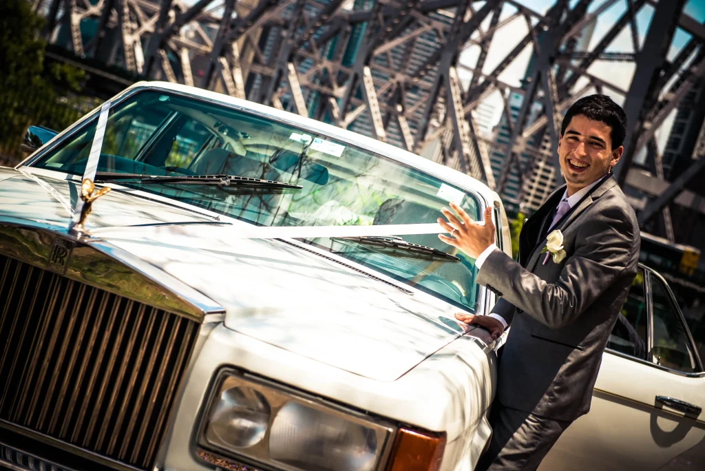 The groom smiles beside the wedding car beneath a bridge.