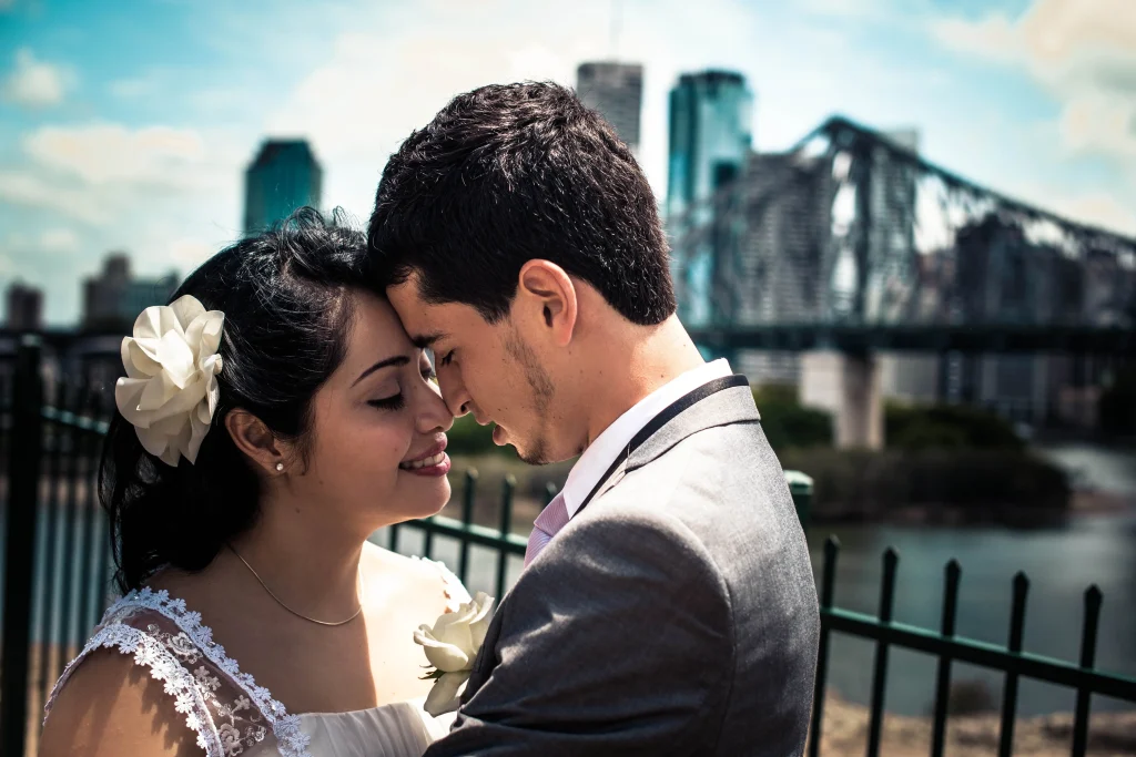 The newlyweds touch foreheads beside the water, with the bridge behind them.